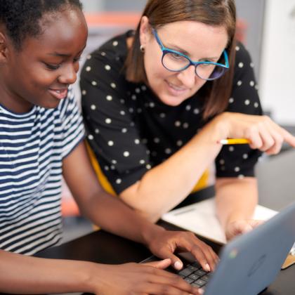 A teacher kneeling beside her student to help her with her computer.