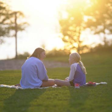 A young girl and camp counsellor talking while sitting on a blanket in the grass.