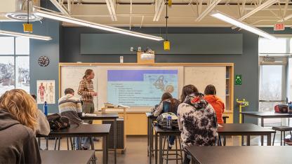 A teacher lecturing to high school students in front of a large learning wall.